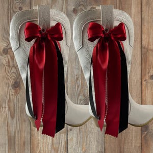 May include: A pair of white cowboy boots featuring decorative attachments on the pull tabs. Each boot is adorned with a large red satin bow, long red and black ribbon streamers, and a delicate strand of white faux pearls hanging vertically.