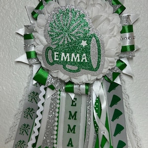 May include: A decorative homecoming mum featuring white silk flowers, green and silver glitter accents, and ribbons. A central cutout displays a green megaphone with the name EMMA in white. Additional ribbons feature printed cheer icons.
