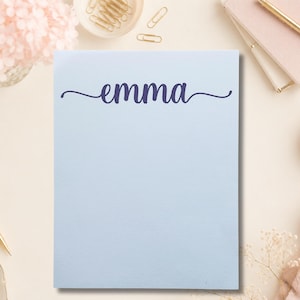 May include: A rectangular light blue notepad featuring the name emma printed in a dark blue cursive script font centered at the top. The paper has a smooth texture and is positioned vertically, displaying a clean and simple design for writing notes.