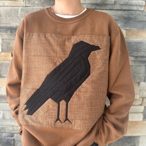 Peut inclure: Un sweat-shirt marron avec un motif de corbeau matelassé noir sur le devant. Le corbeau est bordé de marron et a un fond marron.