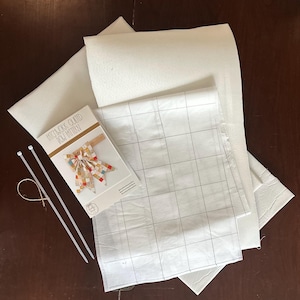 May include: A craft kit containing a Patchwork Quilted Bow Pattern instruction booklet, a sheet of white grid-printed paper, two white plastic zip ties, a piece of twine, and several sheets of white textured material for quilting projects.