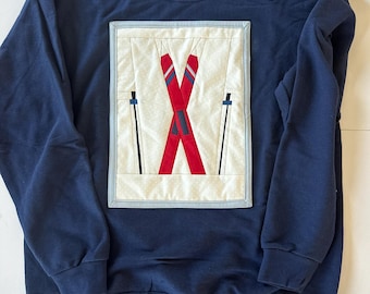 Könnte beinhalten: Ein marineblauer Sweatshirt mit einer weißen, blauen und roten Applikation von gekreuzten Skiern. Die Applikation ist mit einem hellblauen Rand umrahmt.