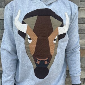 Puede incluir: Una sudadera con capucha gris con un diseño geométrico de cabeza de búfalo marrón, beige y blanco en la parte delantera.
