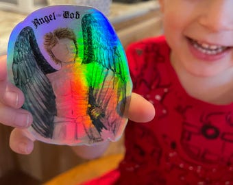 May include: A holographic sticker featuring an angel with outstretched wings. The sticker has the words "Angel of God" at the top. The angel's wings are a mix of colors, including blue, green, and yellow. The angel is wearing a white robe.