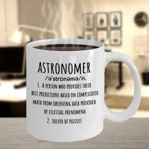 May include: White ceramic mug filled with dark liquid, featuring the word "ASTRONOMER" in black text, along with a definition. The mug has a curved handle and is set against a blurred background.