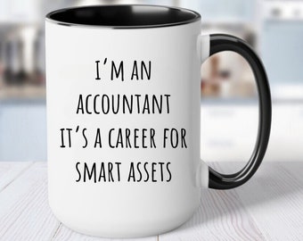 May include: White ceramic mug with a black interior and handle. The mug features the text "I'M AN ACCOUNTANT IT'S A CAREER FOR SMART ASSETS" in black, centered on the front. The mug is on a white wooden surface.