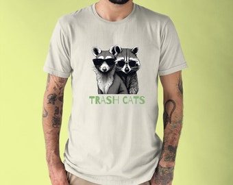 May include: A white t-shirt featuring a black and white illustration of two raccoons wearing sunglasses. The text "TRASH CATS" is printed below the raccoons.