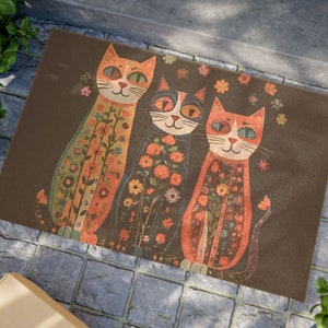 May include: A doormat with three cats wearing floral patterned clothing. The cats are orange, black, and red. The doormat is brown.