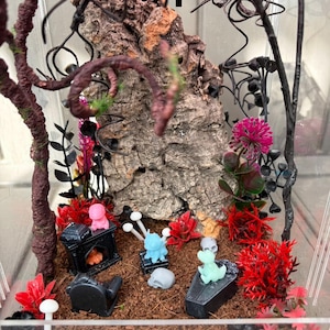 May include: A clear acrylic terrarium containing a large piece of textured brown cork bark, dark brown twisted branches, and red plastic foliage. Miniature items include a black fireplace, a black coffin, a black chair, and small colourful figurines.