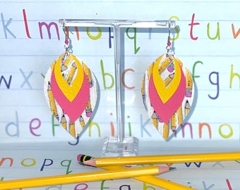 May include: A pair of layered dangle earrings. The earrings feature a white base with a pencil pattern, topped with a pink and yellow layer. The earrings are displayed on a clear stand, with yellow pencils in the foreground.