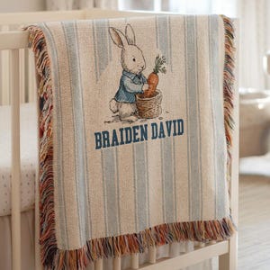 May include: A personalized baby blanket with a bunny illustration holding a carrot. The blanket features blue and white stripes, a fringed edge, and the name "BRAIDEN DAVID" in blue lettering. The blanket is draped over a white crib.