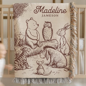 Puede incluir: Una manta beige con una ilustración marrón de Winnie the Pooh y sus amigos, incluyendo un oso, un búho, un conejo y un tigre. La manta tiene un borde con flecos y el nombre "Madeline Jameson" está impreso en ella. La manta está sobre una cuna de madera.