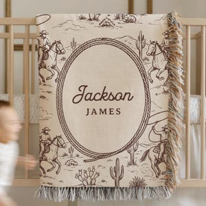 May include: A beige and brown personalised baby blanket with a cowboy theme. The blanket features a desert landscape with cowboys on horseback and cacti. The name "Jackson JAMES" is displayed in a rope-framed oval. The blanket has a fringed edge.