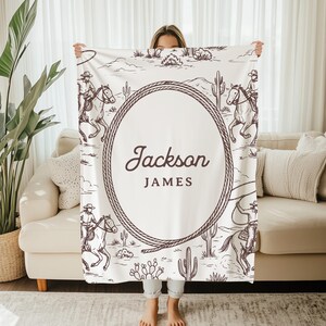 May include: A white blanket with a western design, showing cowboys on horses, cacti, and a rope border. The blanket has the name "Jackson James" in an oval in the centre. The blanket is held up by a person.