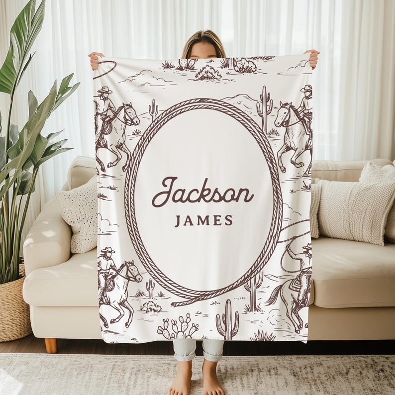 May include: A white blanket with a western design, showing cowboys on horses, cacti, and a rope border. The blanket has the name "Jackson James" in an oval in the centre. The blanket is held up by a person.