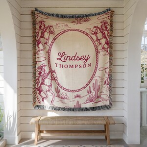 May include: A beige and burgundy woven throw blanket with a western theme, showing cowboys on horseback, cacti, and the text "Lindsey Thompson" within a rope-style oval. The blanket has a fringed edge.