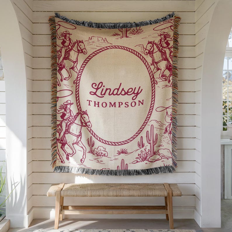 May include: A beige and burgundy woven throw blanket with a western theme, showing cowboys on horseback, cacti, and the text "Lindsey Thompson" within a rope-style oval. The blanket has a fringed edge.