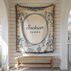 May include: A beige throw blanket with a blue rope design and the name "Jackson JAMES" in blue script. The blanket features a desert scene with cowboys on horseback and cacti. The blanket has a fringed edge and is hanging on a white wall.