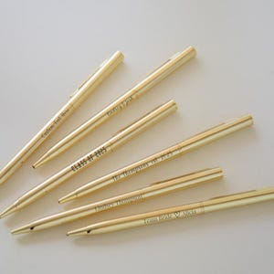 May include: A collection of gold-colored pens with personalized text. The pens have various inscriptions, including "Custom Text Here," "Class of 2025," "Lindsey & Josh," "The Thompsons est. 2025," "Lindsey Thompson," and "Team Bride ♡ Alicia."