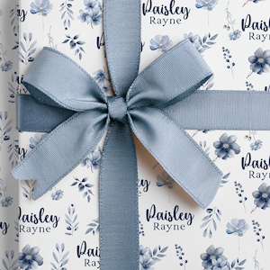 May include: A gift wrapped in white paper with a blue floral pattern and a matching blue ribbon tied in a bow. The paper is decorated with blue flowers and the text "Paisley Rayne".