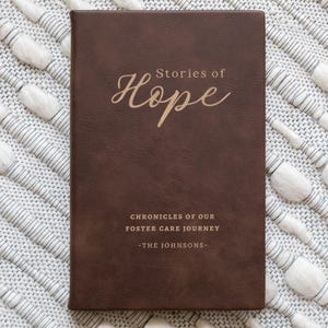 May include: A brown leather-bound journal with the words "Stories of Hope" in gold script. Below, the text reads "Chronicles of Our Foster Care Journey - The Johnsons." The journal is rectangular and lies on a textured, off-white surface.
