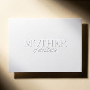 May include: A white card with the embossed text "MOTHER of the Bride" in a serif font. The card is rectangular and sits on a light beige surface, casting a shadow.