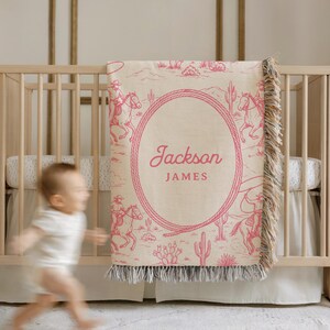 May include: A beige and pink baby blanket with a cowboy design. The blanket depicts a desert scene with cowboys on horseback and cacti. The name "Jackson JAMES" is displayed in a pink oval. The blanket has a fringed edge.