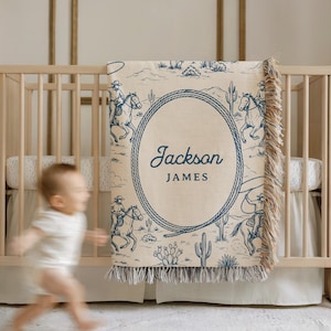 May include: A beige and blue personalised baby blanket with a cowboy theme. The blanket features a desert landscape with cowboys on horseback and an oval frame with the name "Jackson JAMES". The blanket has a fringed edge.