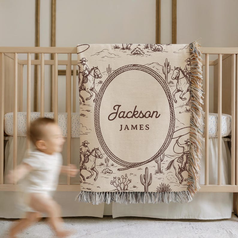 May include: A beige and brown personalised baby blanket with a cowboy theme. The blanket features a desert landscape with cowboys on horseback and cacti. The name "Jackson JAMES" is displayed in a rope-framed oval. The blanket has a fringed edge.