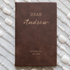 May include: A brown leather journal with the words "DEAR Andrew" and "LETTERS TO MY SON" embossed in gold lettering. The journal is rectangular and lies on a textured white and gray surface.