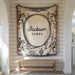 May include: A beige throw blanket with a black and white cowboy design. The blanket features a large oval rope design with the names "Jackson JAMES" in black script. The blanket has a fringed edge and is hanging on a white wall.