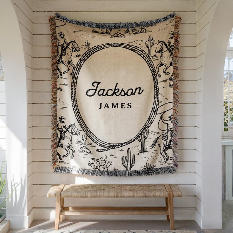 May include: A beige throw blanket with a black and white cowboy design. The blanket features a large oval rope design with the names "Jackson JAMES" in black script. The blanket has a fringed edge and is hanging on a white wall.