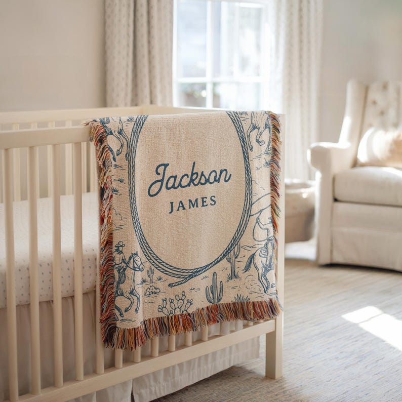 May include: A beige and blue woven baby blanket with a cowboy theme, draped over a white cot. The blanket features a blue oval with the name "Jackson JAMES" and a design of cowboys, horses, and cacti. The blanket has a fringed edge.