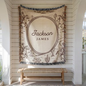 May include: A beige woven throw blanket with a western theme, featuring cowboys on horseback, cacti, and a rope design. The blanket has the name "Jackson JAMES" in a brown script font. The edges are fringed.