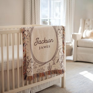 May include: A beige and brown personalised baby blanket with a cowboy theme. The blanket features a rope border with the name "Jackson JAMES" in the centre. The design includes illustrations of cowboys, horses, and cacti. The blanket has a fringed edge.