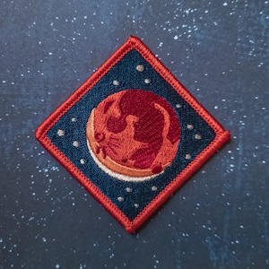 May include: A red and orange embroidered patch of a crescent moon with a red planet in the background. The patch is on a dark blue background with white stars. The patch is diamond shaped with a red border.