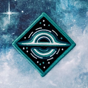 May include: A teal blue diamond-shaped patch with a black background and a white and teal blue embroidered design of a black hole with a white ring around it.