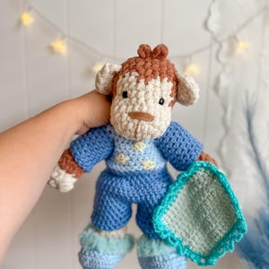 Peut inclure: Poup&eacute;e singe crochet&eacute;e portant un pyjama bleu et des chaussons bleus duveteux. La poup&eacute;e a des d&eacute;tails marron et cr&egrave;me, notamment le visage, les oreilles et les cheveux. Elle tient une petite couverture carr&eacute;e avec une bordure turquoise.