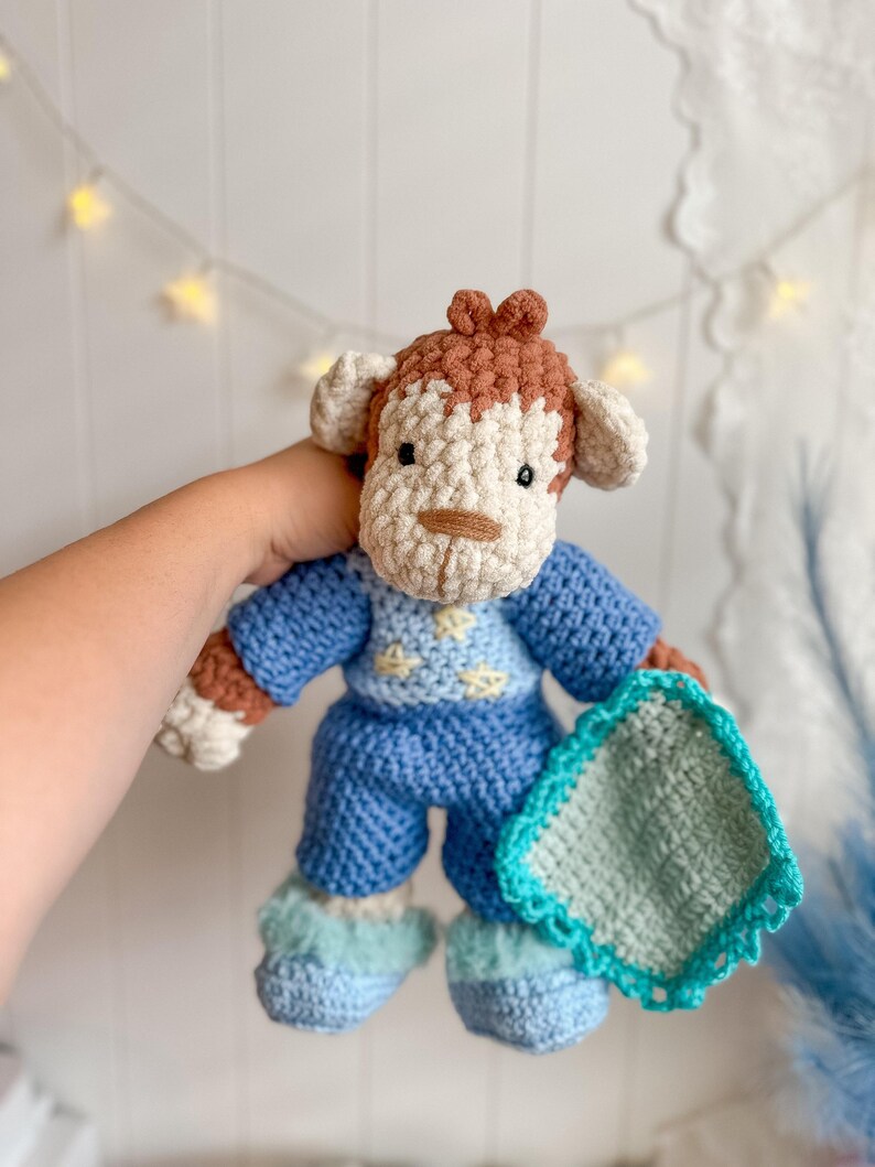 Peut inclure: Poup&eacute;e singe crochet&eacute;e portant un pyjama bleu et des chaussons bleus duveteux. La poup&eacute;e a des d&eacute;tails marron et cr&egrave;me, notamment le visage, les oreilles et les cheveux. Elle tient une petite couverture carr&eacute;e avec une bordure turquoise.
