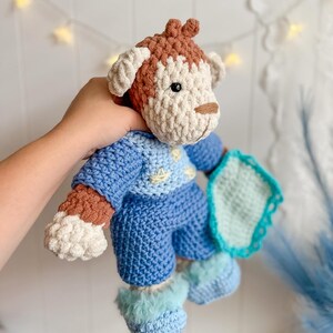 Peut inclure: Une poup&eacute;e chien au crochet faite &agrave; la main dans les tons de bleu, marron et cr&egrave;me. Le chien porte une tenue bleue avec une couverture bleu clair. La poup&eacute;e a une t&ecirc;te marron avec des oreilles et un museau cr&egrave;me. La tenue du chien comprend un pull, un pantalon et des chaussons.