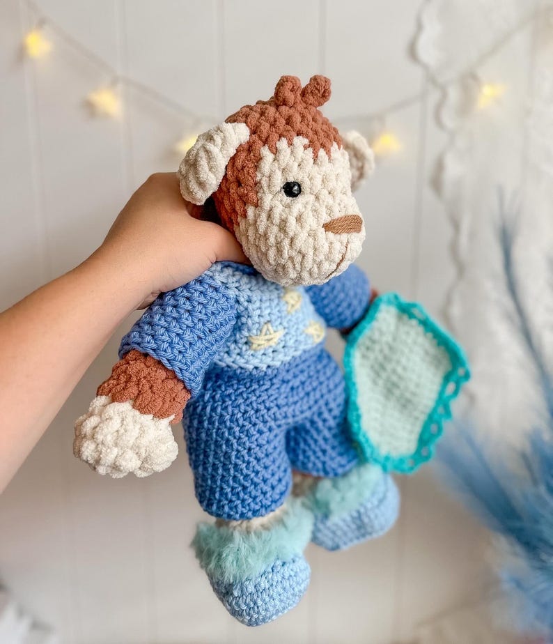 Peut inclure: Une poup&eacute;e chien au crochet faite &agrave; la main dans les tons de bleu, marron et cr&egrave;me. Le chien porte une tenue bleue avec une couverture bleu clair. La poup&eacute;e a une t&ecirc;te marron avec des oreilles et un museau cr&egrave;me. La tenue du chien comprend un pull, un pantalon et des chaussons.
