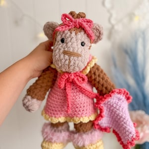 Peut inclure: Poup&eacute;e singe au crochet faite &agrave; la main, v&ecirc;tue d'une robe rose, d'un n&oelig;ud et de chaussures assorties. Le singe a des bras et des jambes marron, un visage beige et un n&oelig;ud rose. La robe est bord&eacute;e de jaune et tient un accessoire rose et rouge.