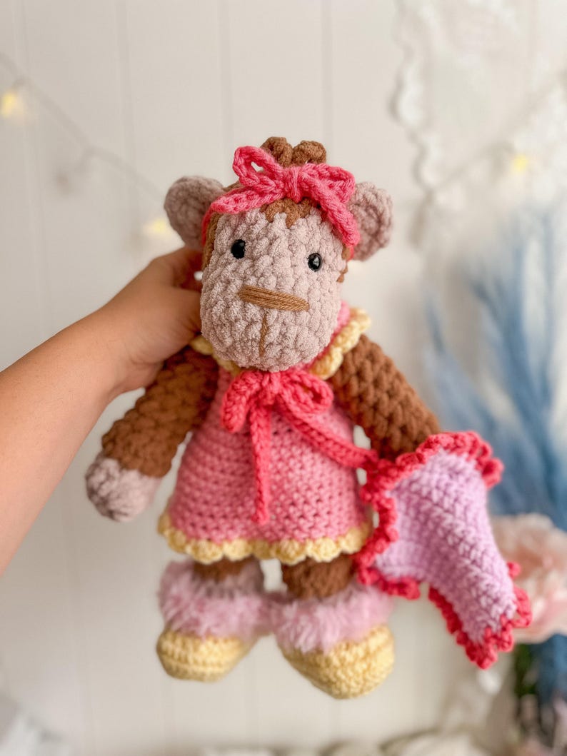 Peut inclure: Poup&eacute;e singe au crochet faite &agrave; la main, v&ecirc;tue d'une robe rose, d'un n&oelig;ud et de chaussures assorties. Le singe a des bras et des jambes marron, un visage beige et un n&oelig;ud rose. La robe est bord&eacute;e de jaune et tient un accessoire rose et rouge.