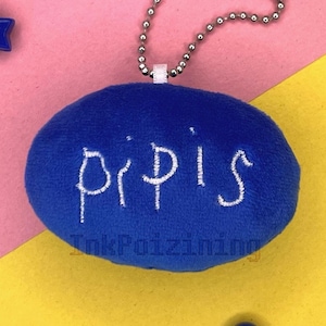 May include: Blue plush keychain with a silver chain and the word "pipis" embroidered in white. The keychain is on a yellow and pink background with blue stars and colorful beads.