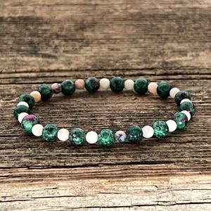 Puede incluir: Un brazalete de cuentas verdes, rosas y blancas. El brazalete está hecho de pequeñas cuentas redondas y está enhebrado en un cordón elástico transparente.