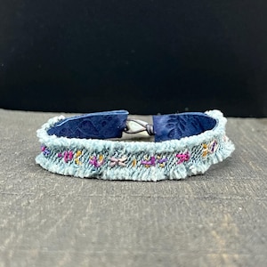 Puede incluir: Un brazalete de mezclilla azul con cierre de cuero y detalles florales bordados en hilo rosa, morado y dorado.