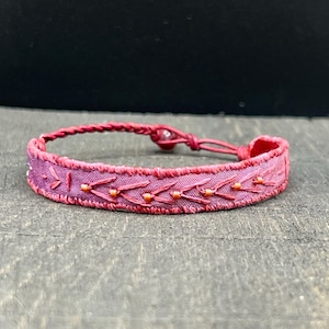 May include: A handmade bracelet with a maroon fabric band and intricate embroidery. The band features a repeating pattern of embroidered arrows and small orange beads. The adjustable closure is a braided cord. This bracelet is a unique piece of jewelry.