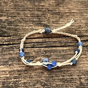 May include: A white braided cord bracelet with blue gemstone beads. The bracelet is tied with a knot at the end.