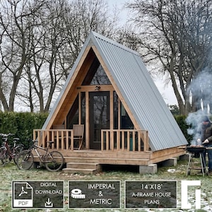 May include: A digital download of 14'x18' A-frame house plans. The structure features a wooden frame, a gray corrugated metal roof, a front porch with wooden railings, and a central door. Three bicycles are parked on the grass nearby.