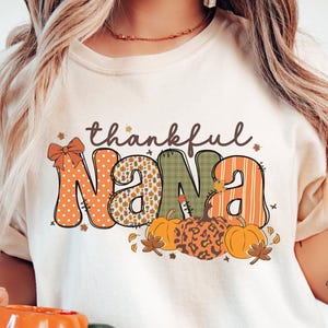 May include: Cream-colored t-shirt with the words "thankful Nana" in a fall-themed design. The word "Nana" is decorated with orange polka dots, green plaid, and orange stripes. Pumpkins and leaves are at the bottom.